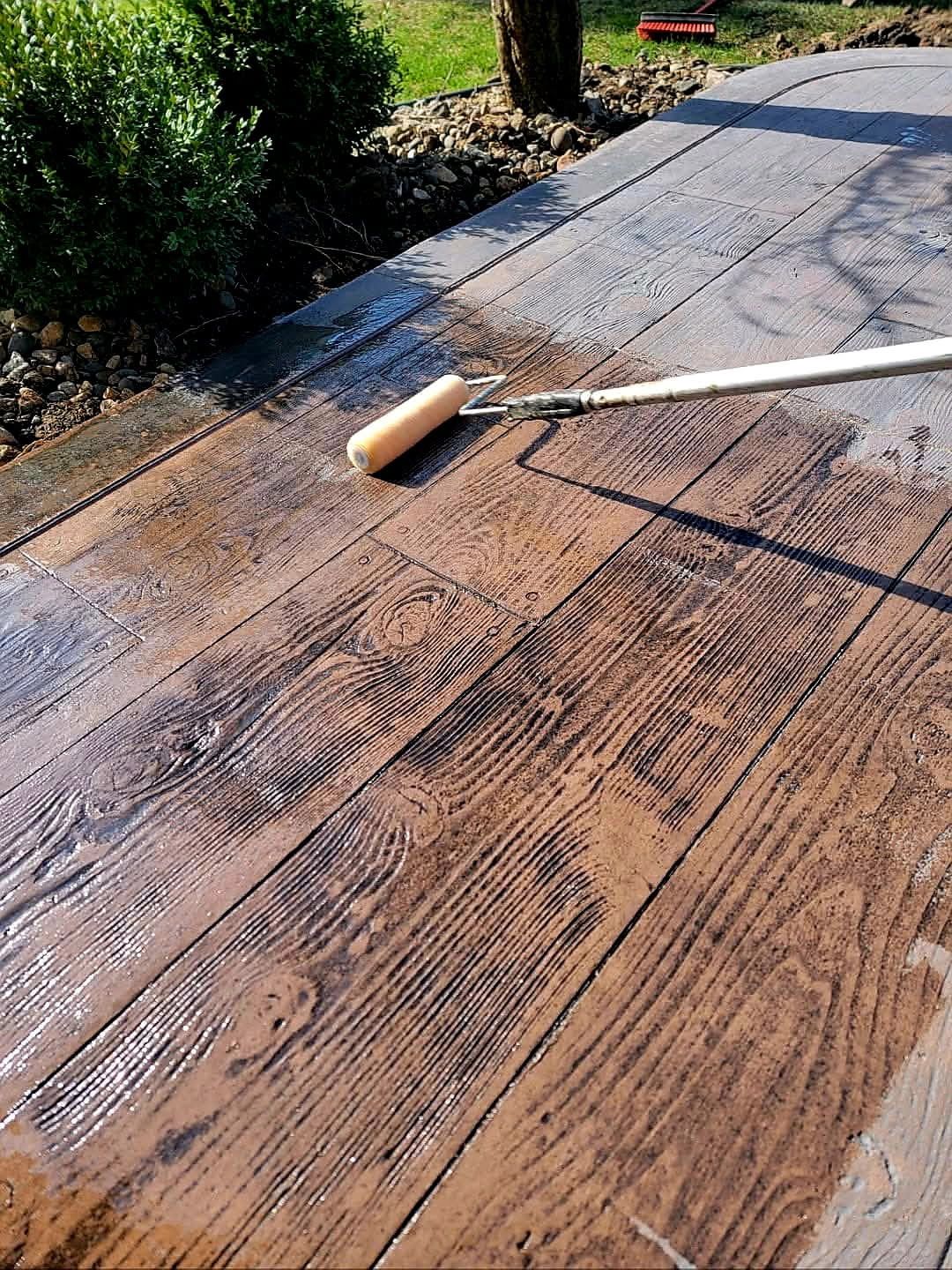 A person using a paint roller applies sealant to stamped concrete patio, wood grain pattern.