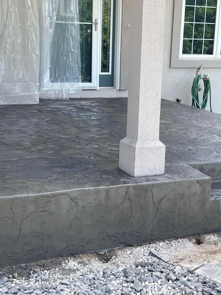 Grey concrete porch with pillar, steps, and door. Building under construction, surrounded by gravel.