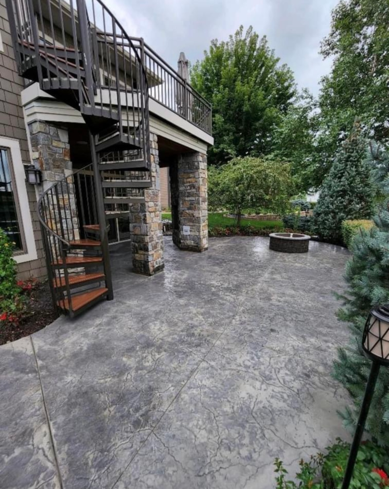 Spiral staircase and patio with fire pit, stone columns, and surrounding greenery.