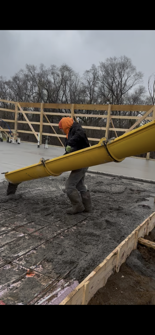 Construction worker pouring concrete from a yellow chute into a form, trees in the background, overcast sky.