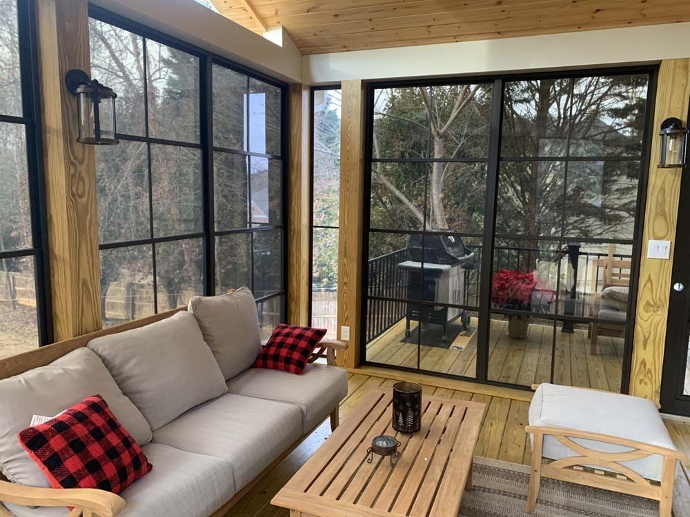 Sunroom with a beige sofa, wooden table, and large black-framed windows overlooking a wooded area.