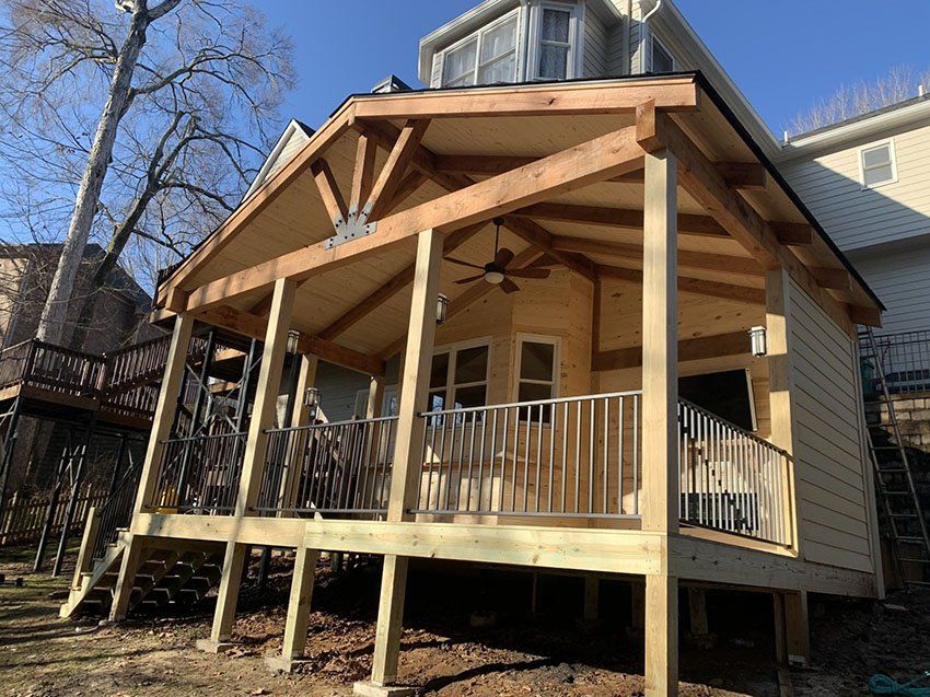 A newly built wooden porch with a gabled roof, connected to a beige-sided house, with a railing and a ceiling fan.