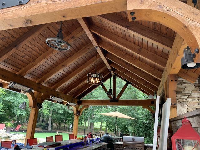 Wooden outdoor pavilion with a bar area, ceiling fans, and an open view of a backyard with a pool and trees.