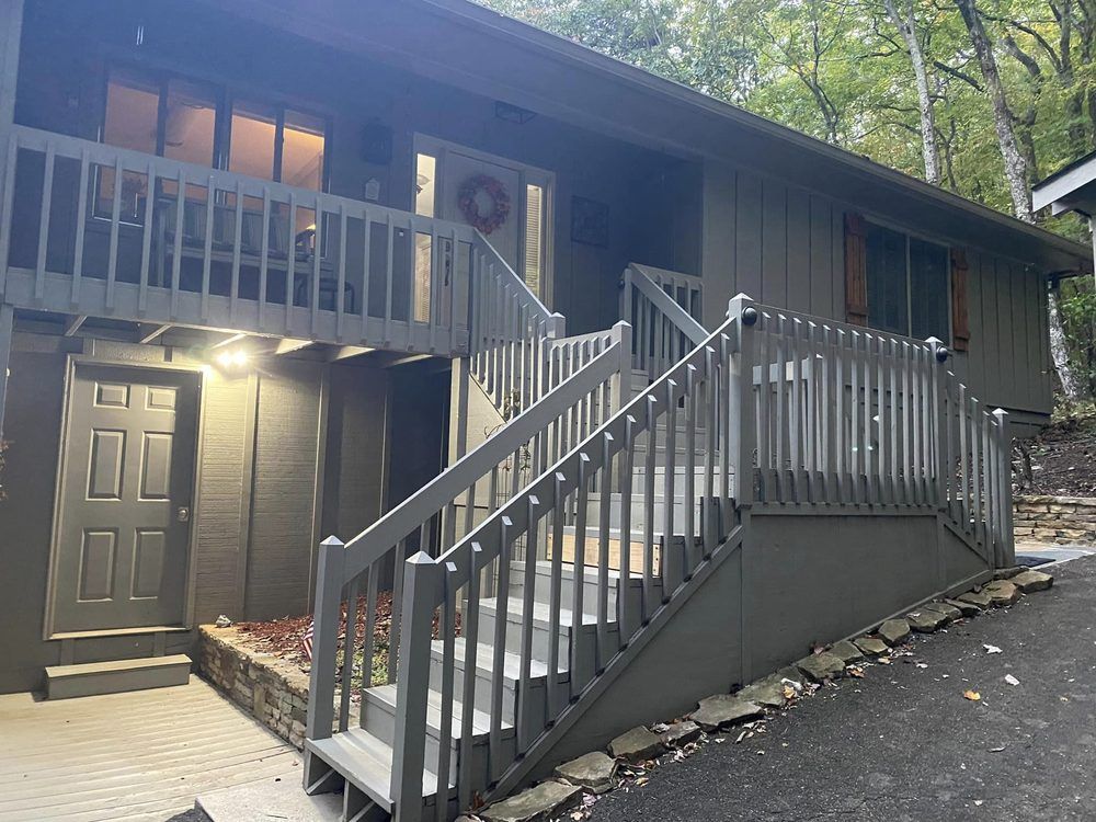 A gray two-story house with a front staircase, walkway, and porch, set against a forest background.