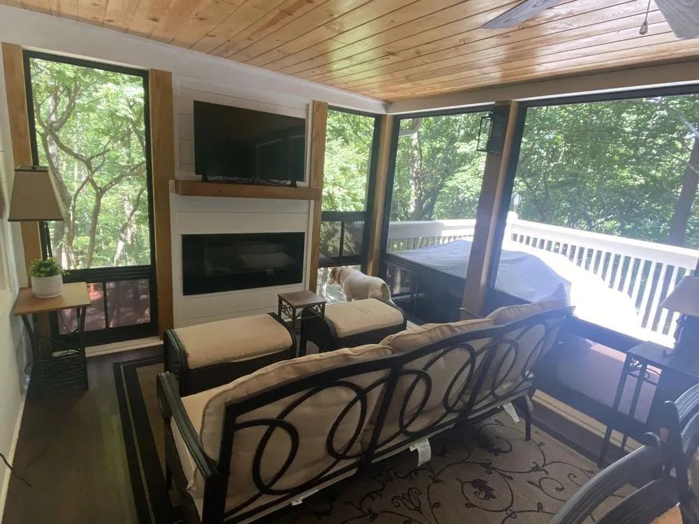 Screened-in porch with sofa, fireplace, and TV overlooking trees.