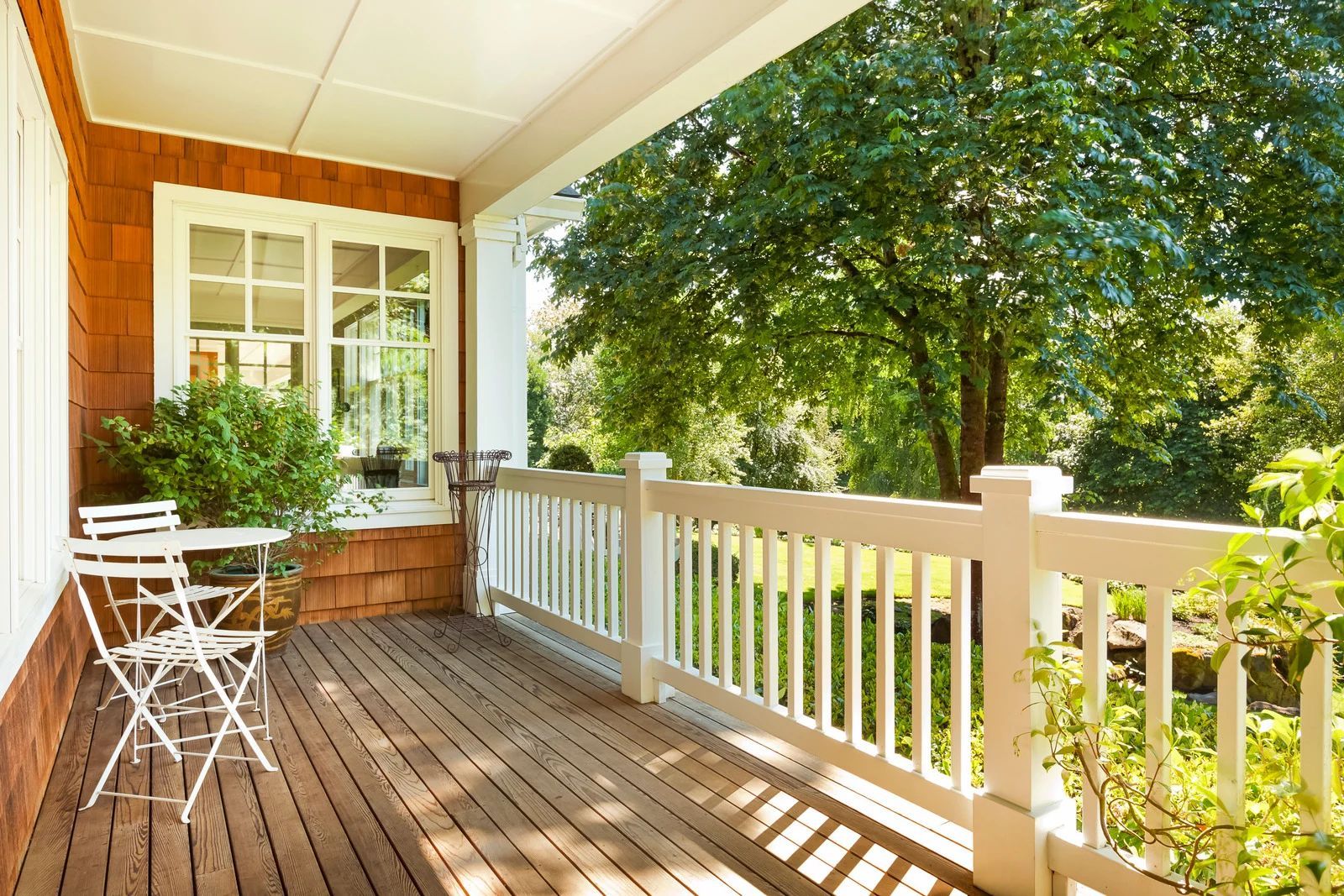 White porch with wooden floor, small table and chairs, and a view of green trees beyond the railing.