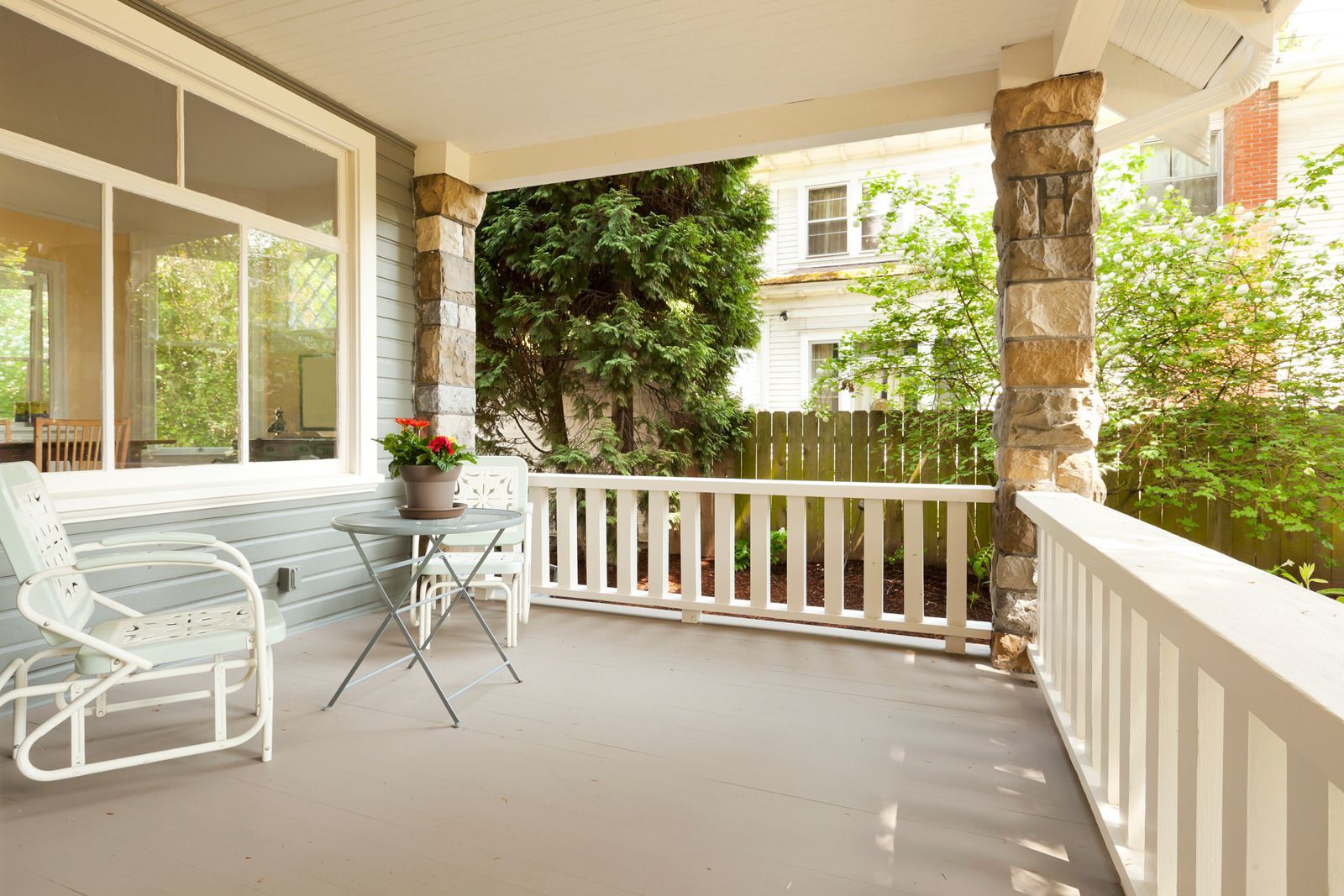 Covered porch with white railing, wicker chair, small table, and lush greenery outside