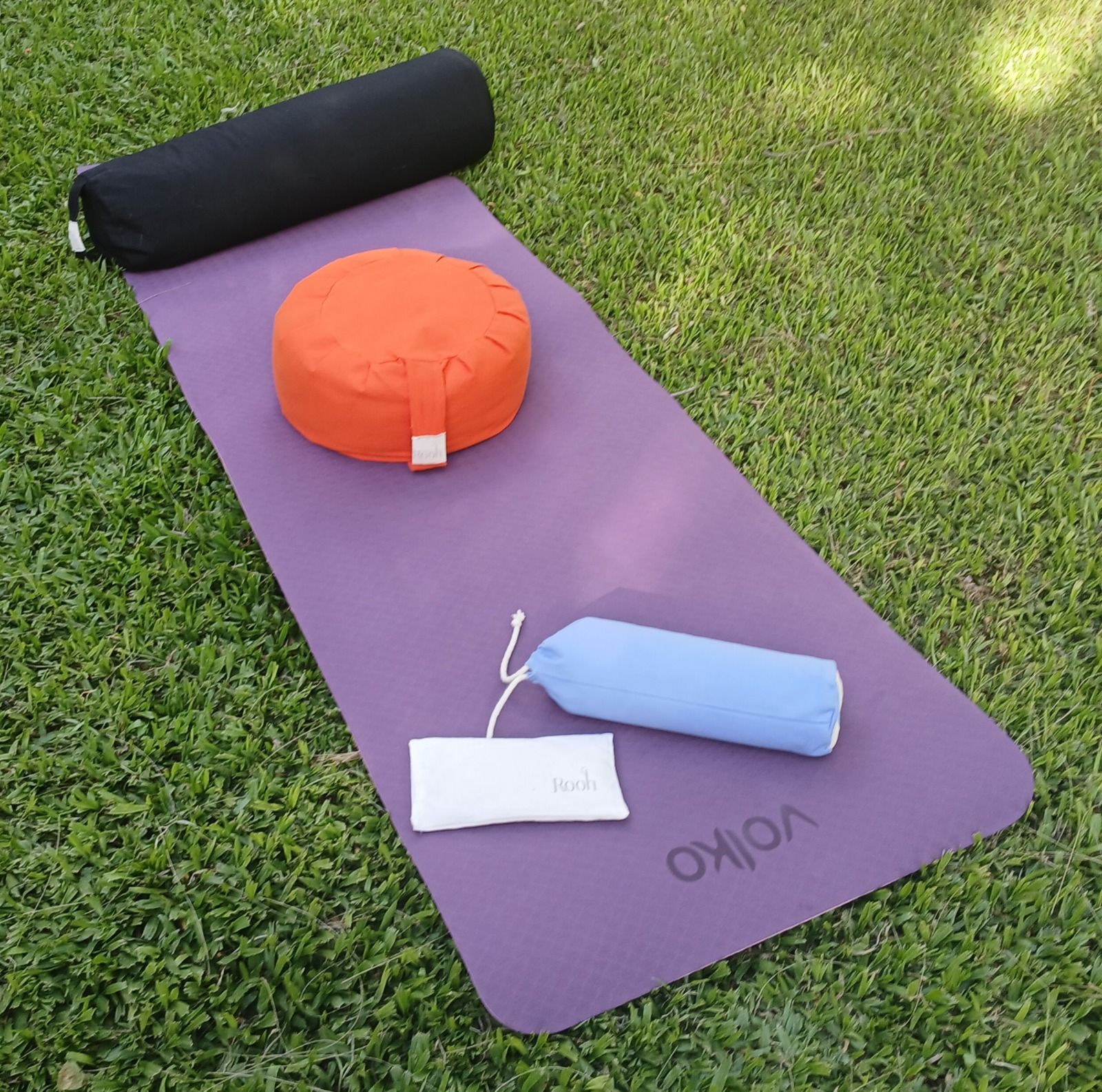 Esterilla de yoga sobre césped con un almohadón negro, un cojín naranja, un cilindro azul y una bolsa con correa blanca.