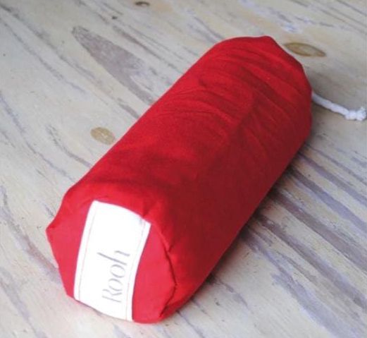 Cojín cilíndrico de tela roja con una etiqueta blanca, sobre un suelo de madera clara.