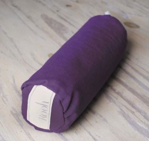 Cojín cilíndrico de tela morada sobre un suelo de madera clara.