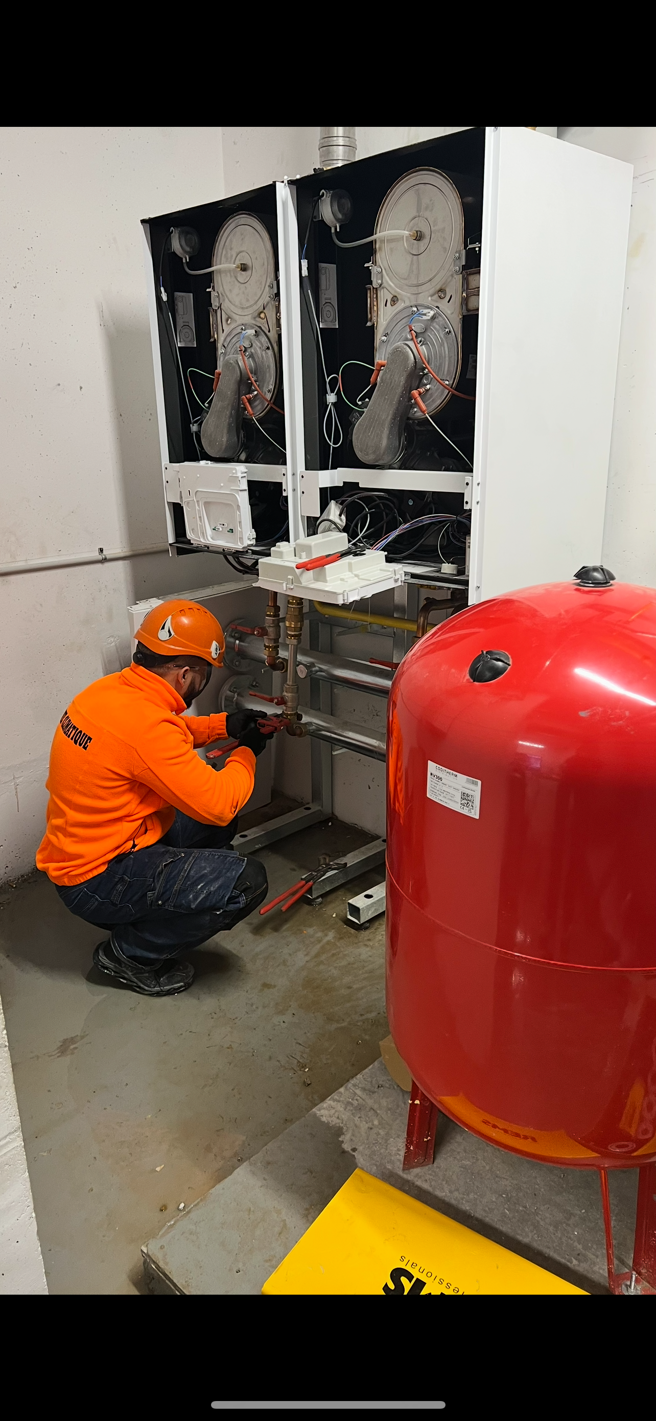 Technicien en uniforme orange travaillant sur un équipement industriel à côté d'un réservoir rouge.