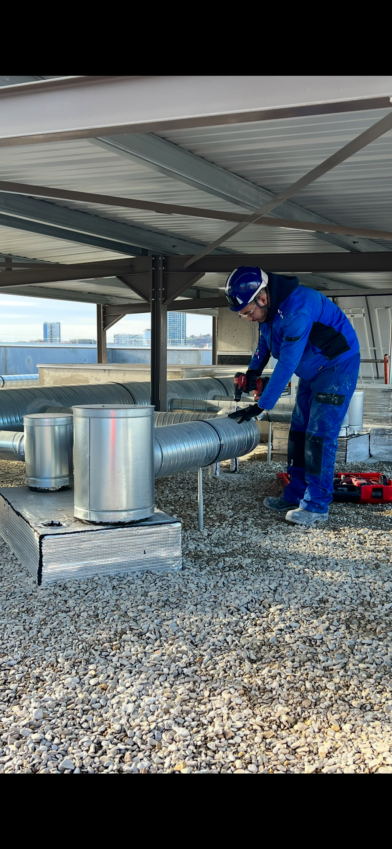 Un ouvrier en uniforme bleu inspecte les conduits de ventilation sur le toit, sous un auvent métallique.