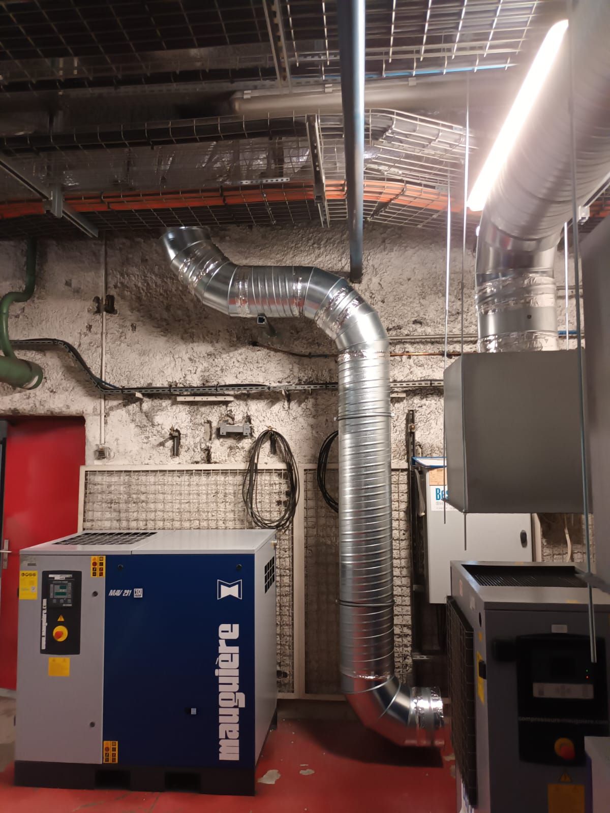 Sous-sol industriel avec compresseur d'air bleu et grands conduits de ventilation argentés adossés à un mur de briques.