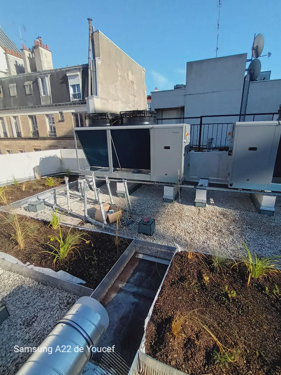 Aire utilitaire sur le toit avec unités de climatisation, gravier et un étroit passage piétonnier entre les bâtiments bas.