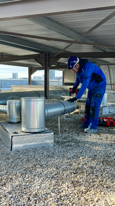 Un ouvrier en uniforme bleu inspecte les conduits de ventilation du toit sous un auvent métallique.