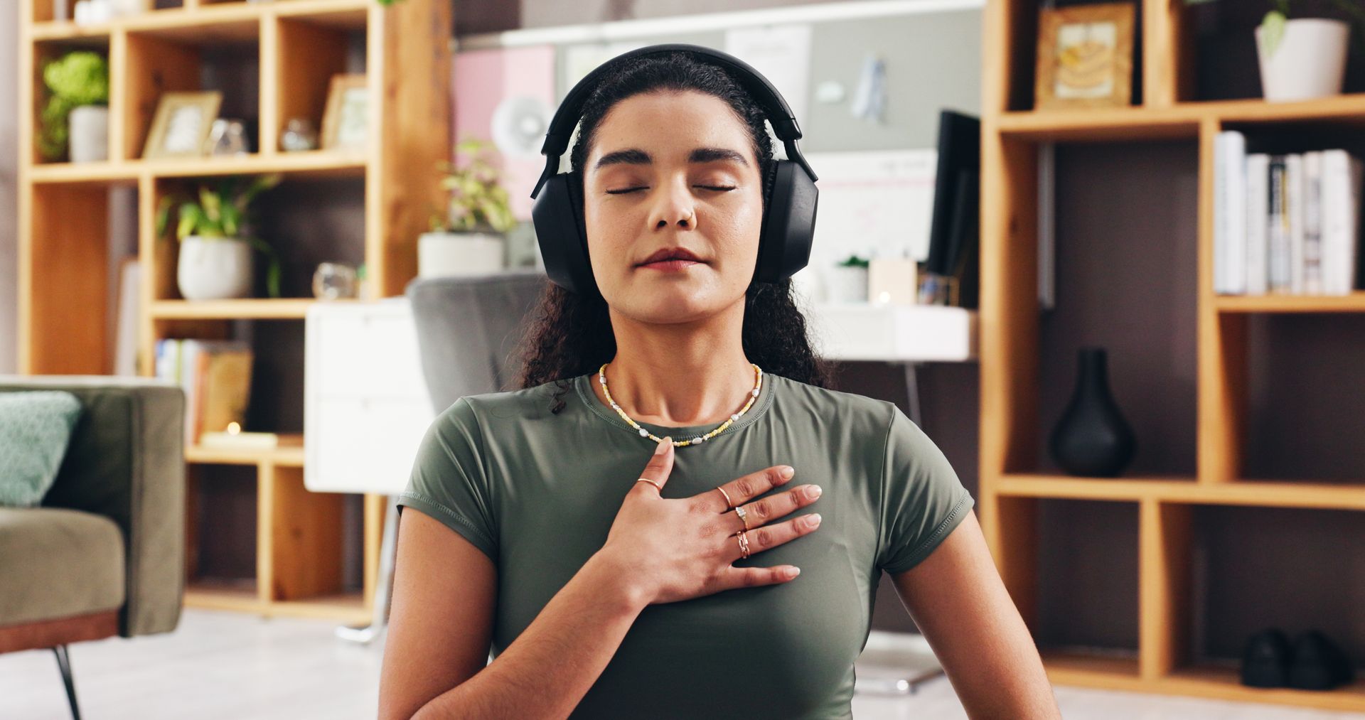 Person meditating indoors with hand on chest and headphones on, in front of bookshelves
