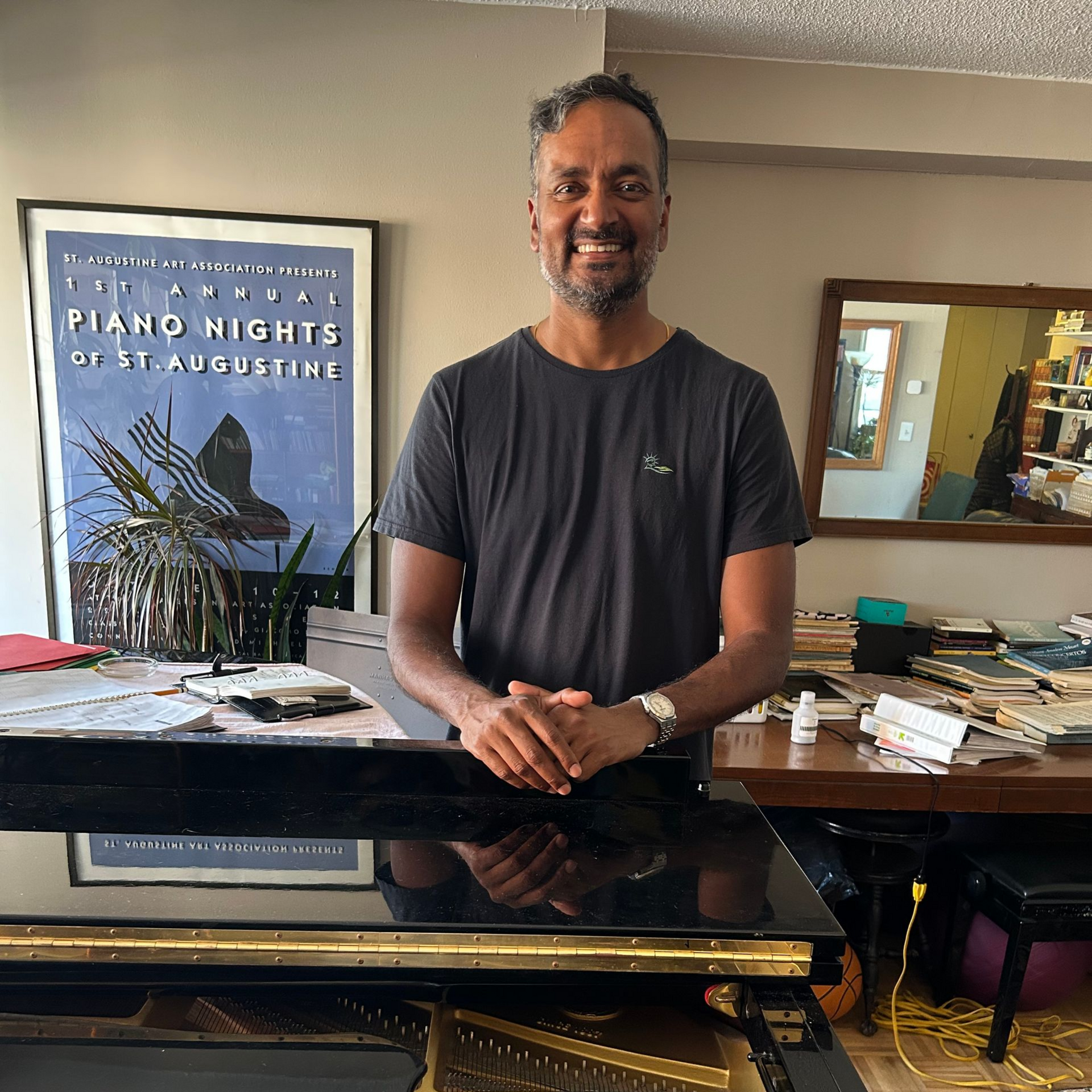 Man smiling behind a glossy black piano in a room with a “Piano Nights” poster.