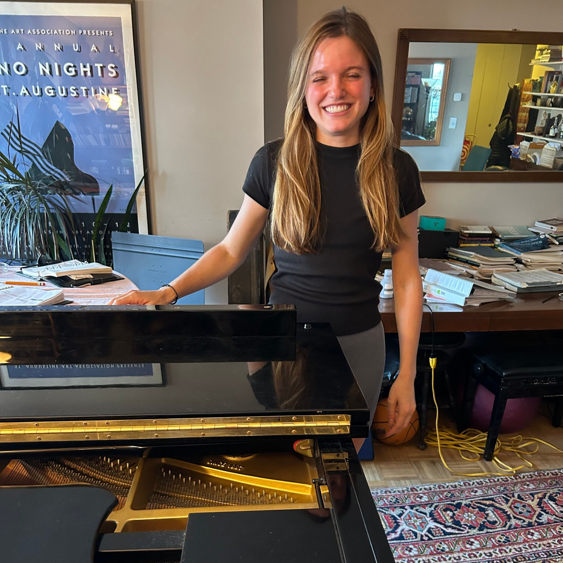 Smiling woman standing behind a black piano in a room with books and a poster.