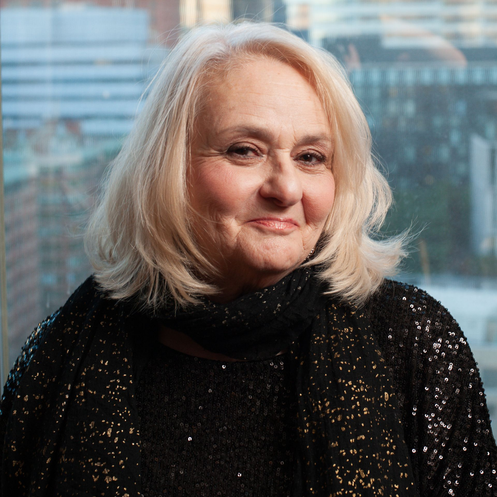 Portrait of a smiling woman with shoulder-length white hair in a black sweater, seated before a city window backdrop