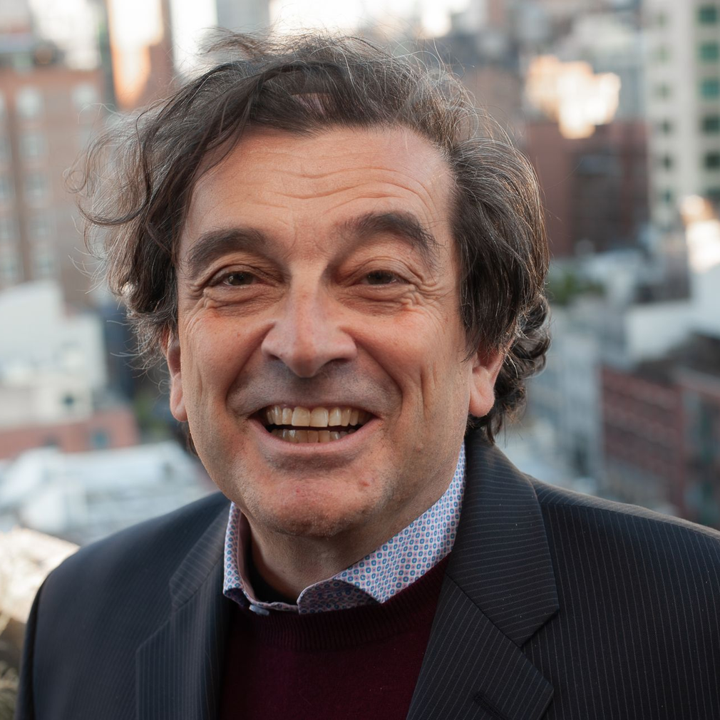 Smiling man in a dark blazer and sweater, standing outdoors with a blurred city background