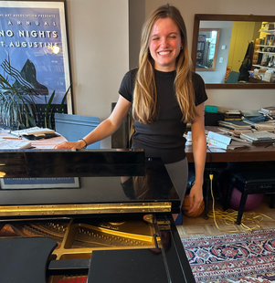 Smiling person stands beside a grand piano in a cozy music room.