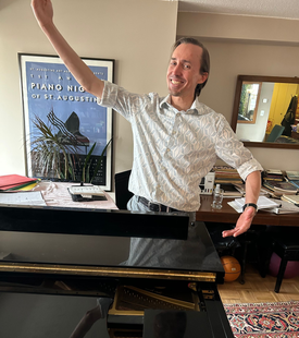 Man smiling and gesturing beside a grand piano in a room with framed artwork and a patterned rug
