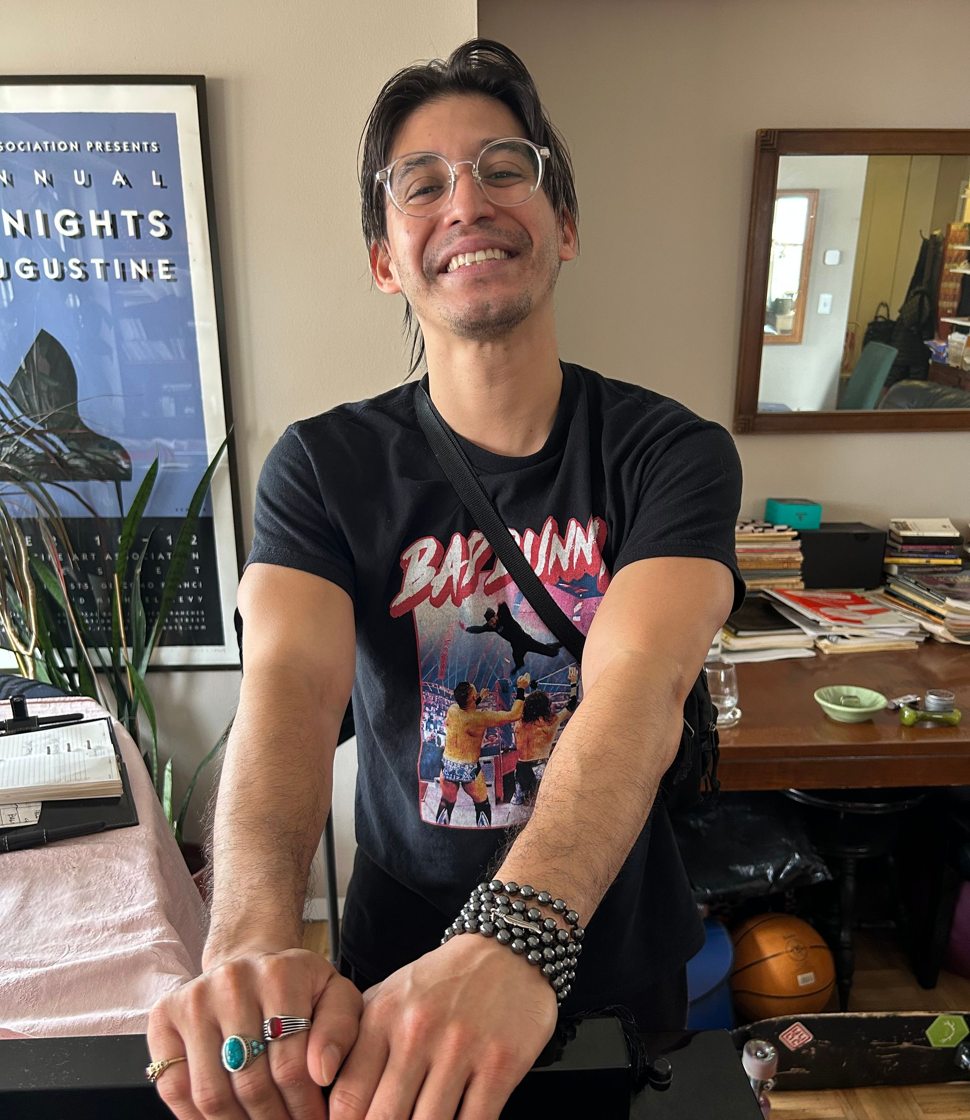 Smiling person in a black graphic T-shirt posing indoors with arms crossed over a desk.