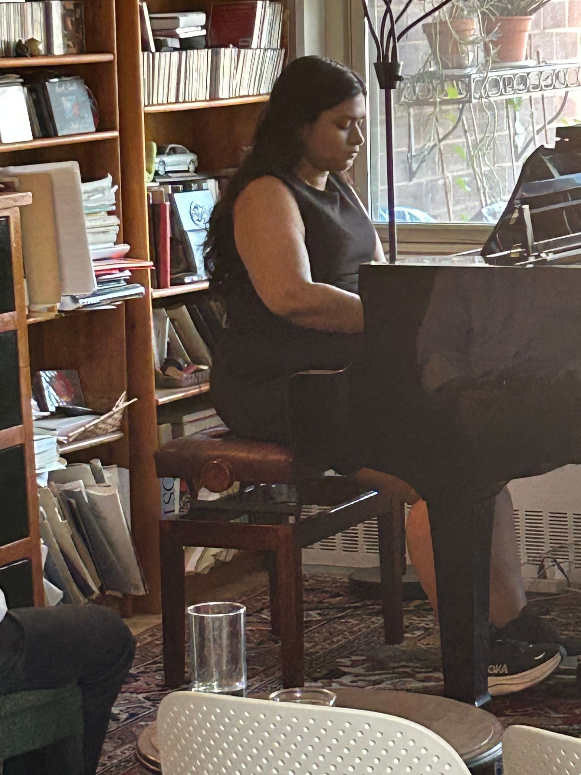 Person playing piano in a cozy café, seated beside shelves and a sunlit window.