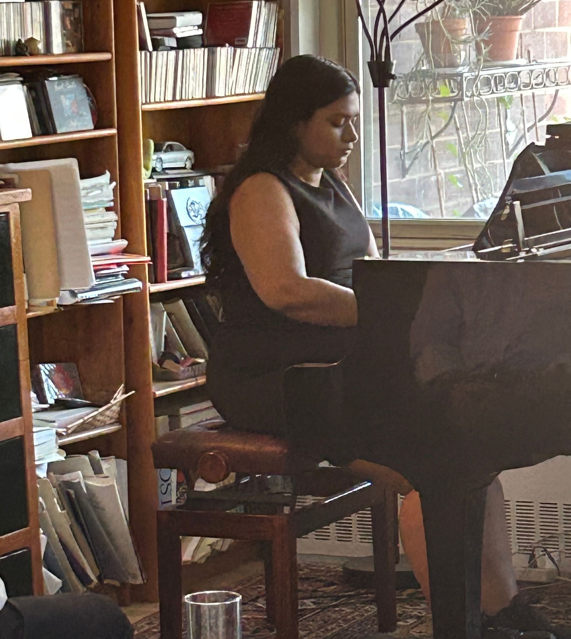 Person seated at a piano in a cozy room, with bookshelves and a window in the background