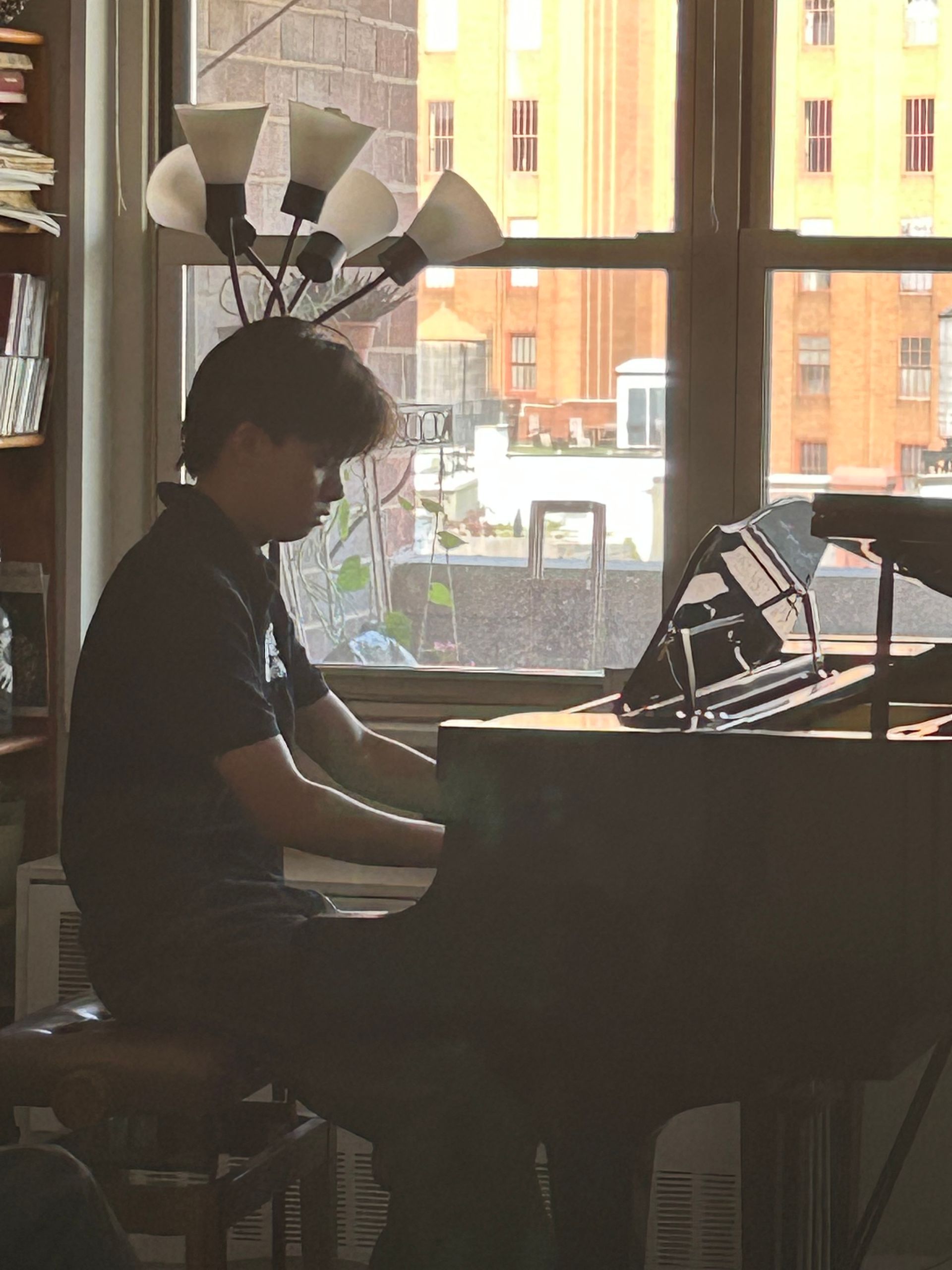 Person playing a piano in a dimly lit room by a large window with city buildings outside