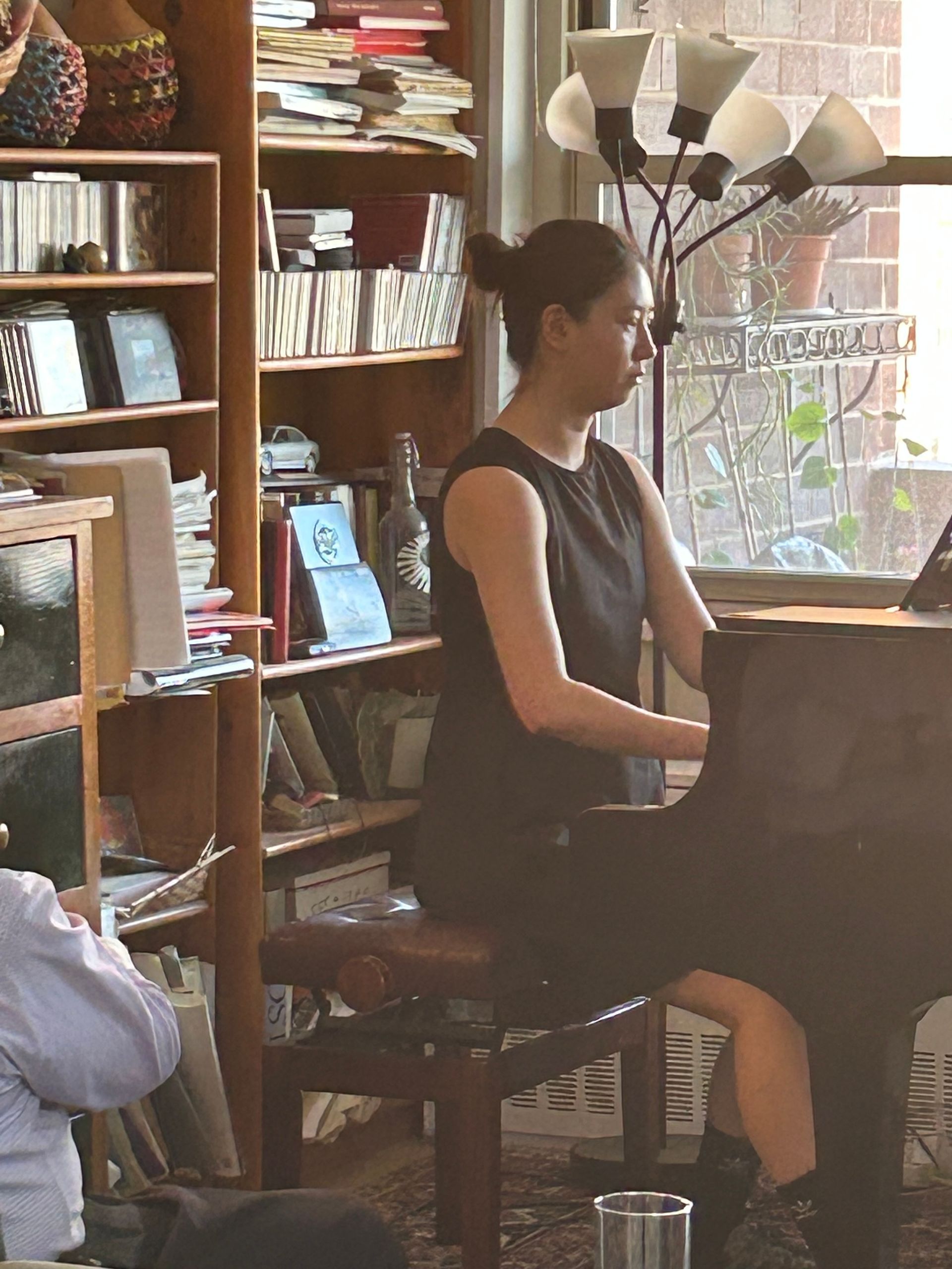 Person seated at a piano in a cozy room with bookshelves and a window.