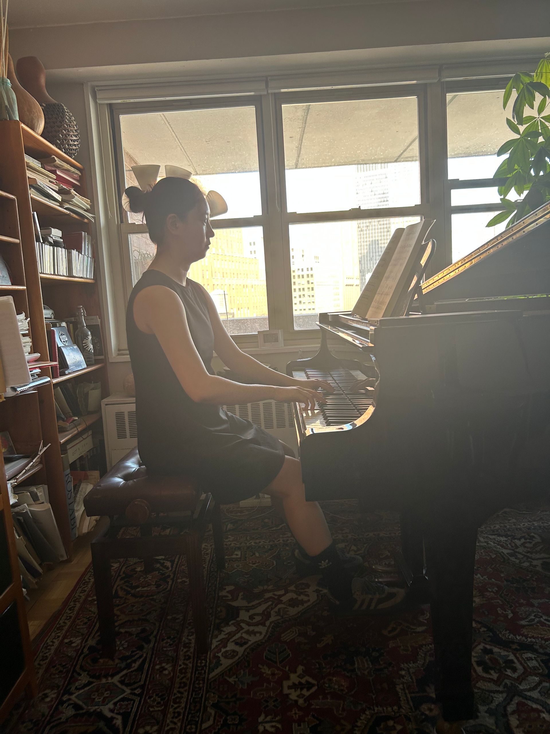 Person playing piano in a sunlit room with bookshelves and a patterned rug
