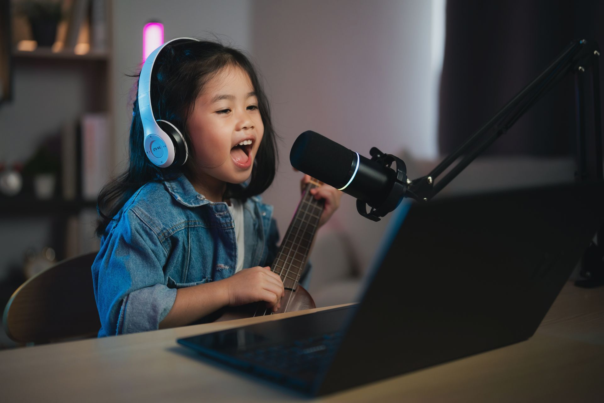 Child wearing blue headphones sings into a microphone while holding a ukulele at a desk with a laptop.