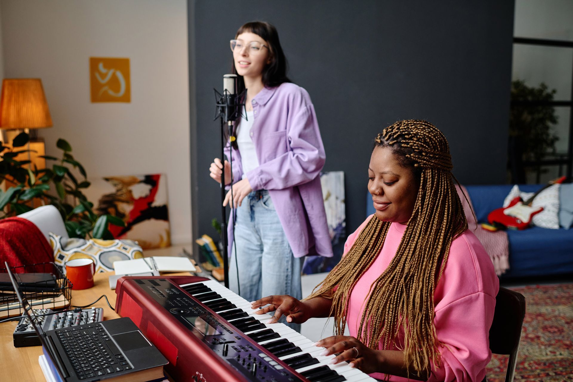 Two women making music in a home studio, one singing into a mic and one playing a keyboard