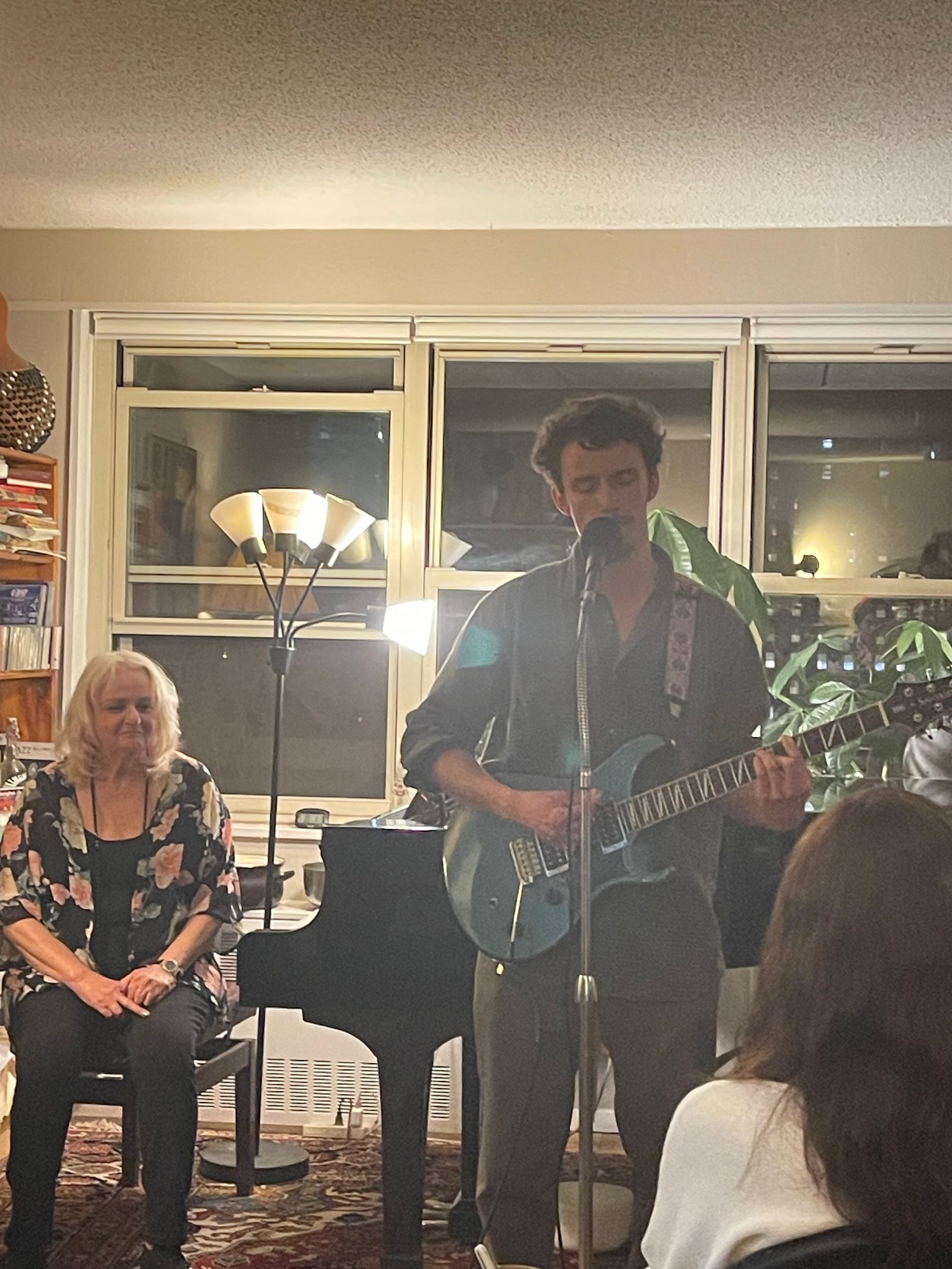 Musician singing and playing guitar at a small indoor gathering, with seated audience and warm lamp lighting.