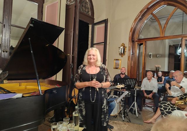 Singer performing beside a grand piano in an elegant room with seated musicians and audience.