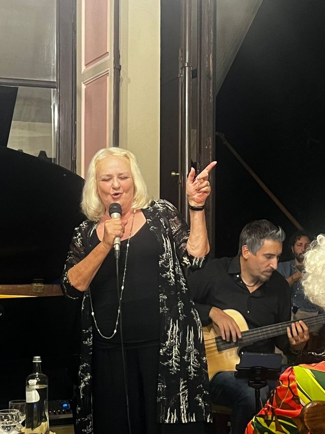 Singer performing with microphone on stage, pointing upward, while a guitarist plays beside her.