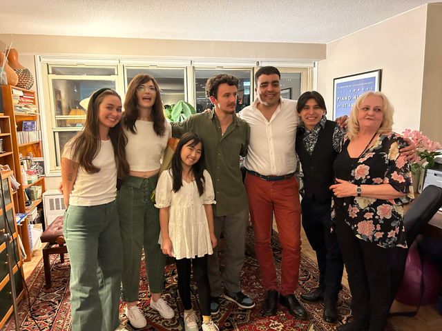 Six people posing together in a living room, smiling in casual clothes with shelves and framed art behind them.