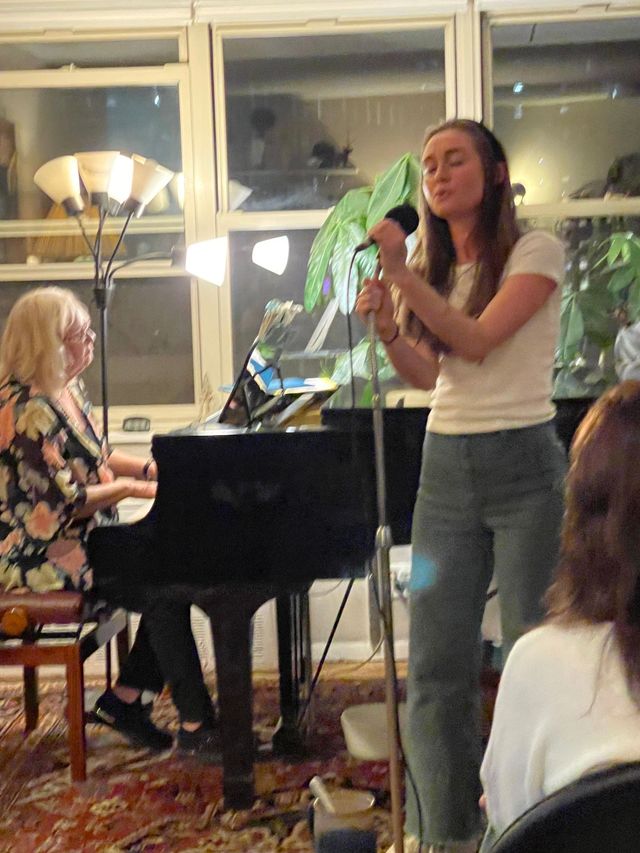 Woman singing beside a piano while a seated pianist plays in a warmly lit room