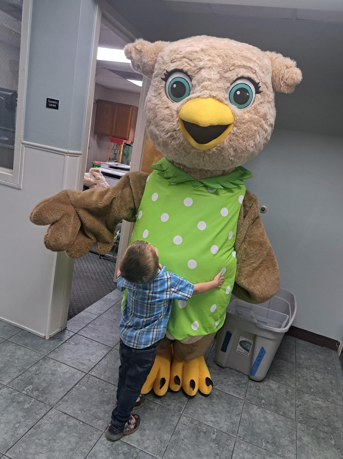 A little boy is hugging an owl mascot in a green dress.