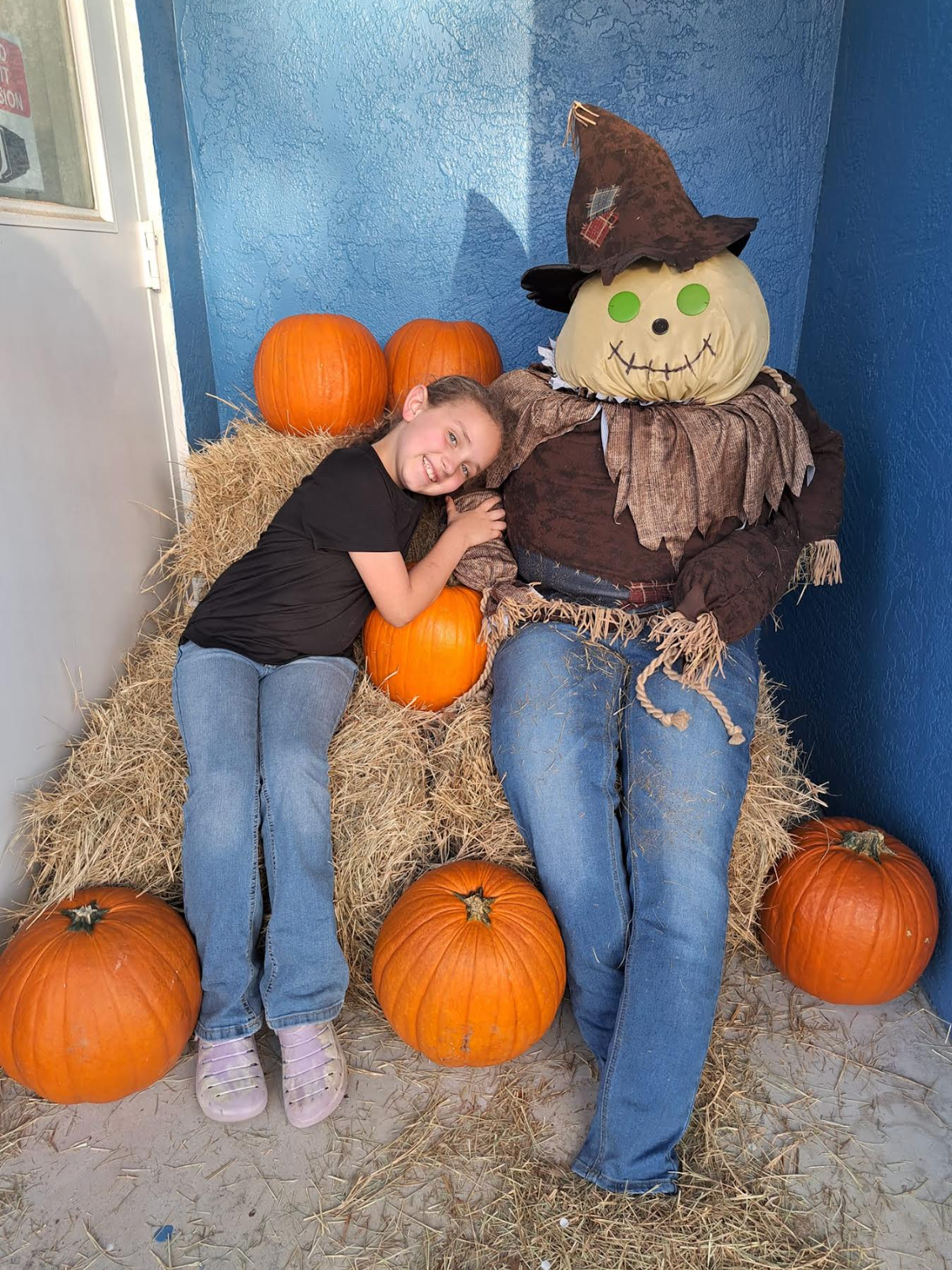 A little girl is laying next to a scarecrow surrounded by pumpkins.