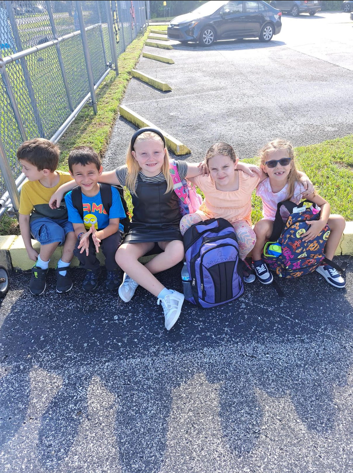 A group of children are sitting on the side of the road with their backpacks.