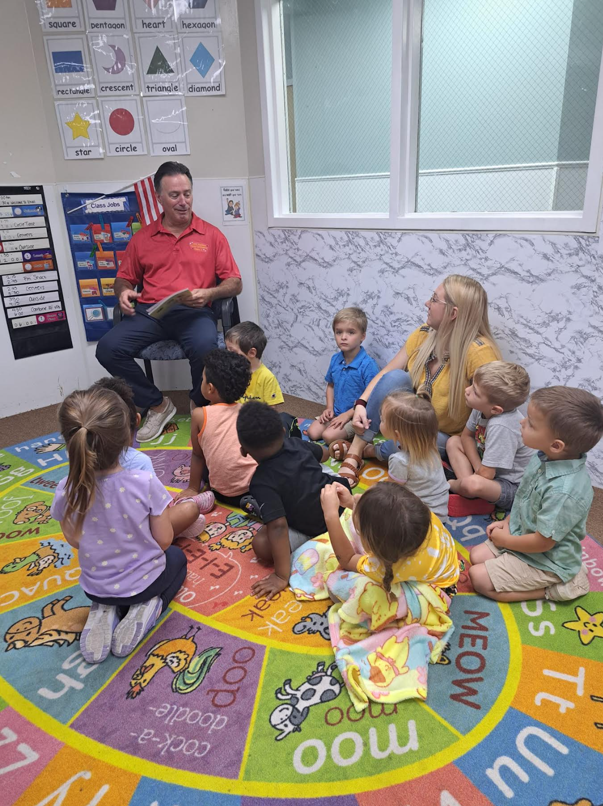 A group of children are sitting on the floor listening to a man read a book.