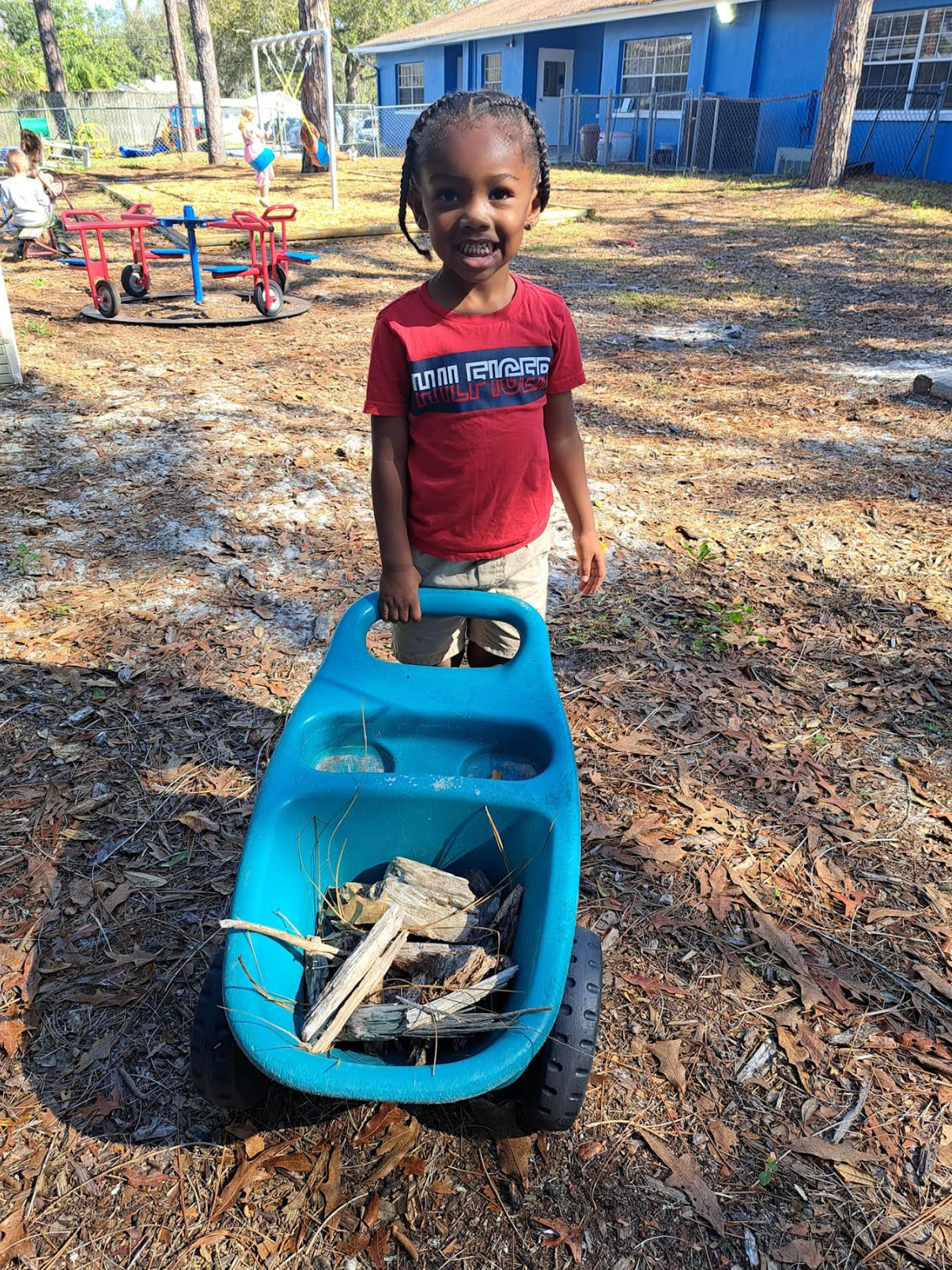 A young boy is standing next to a blue wheelbarrow filled with wood.