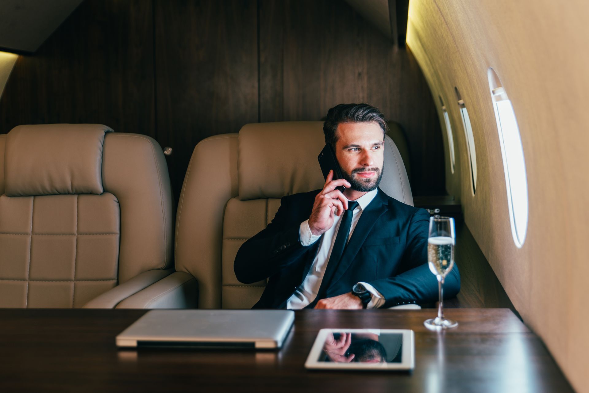 A man in a suit is sitting at a table on a private jet talking on a cell phone.