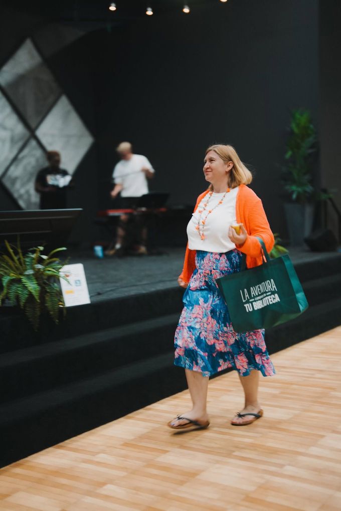 Woman walking indoors with a green tote bag, in a modern event space with plants and steps
