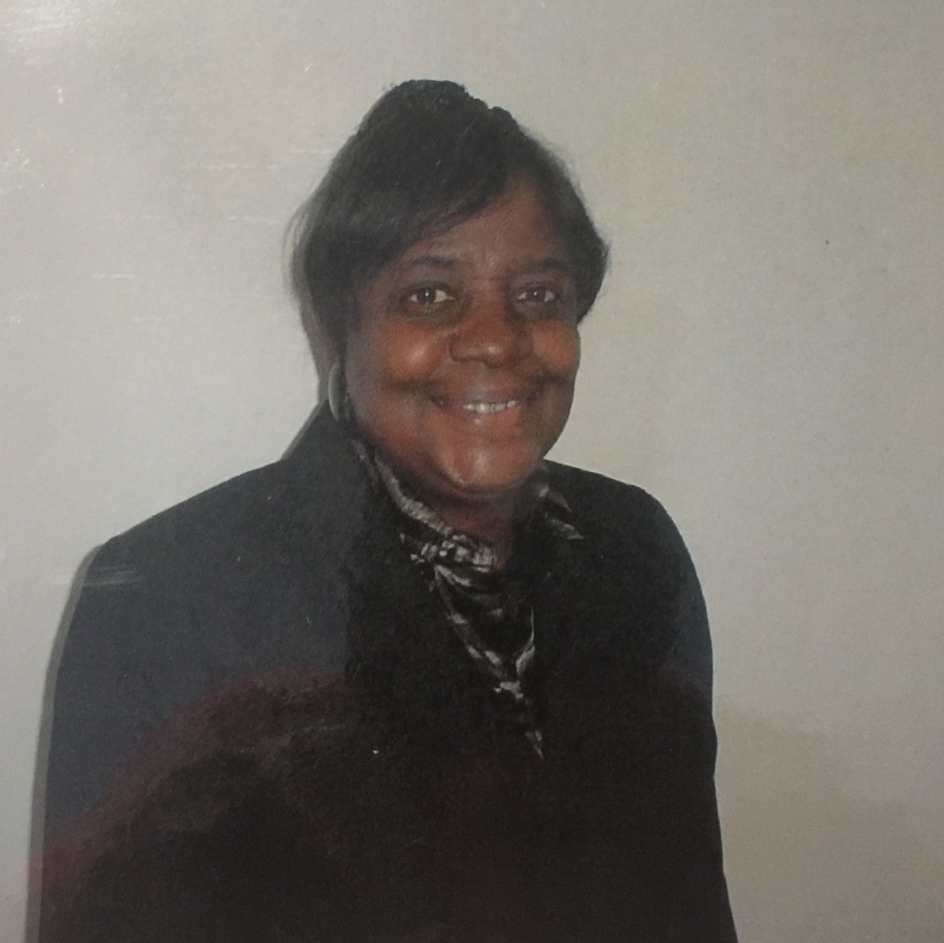 Smiling person in a dark jacket standing against a plain light wall.