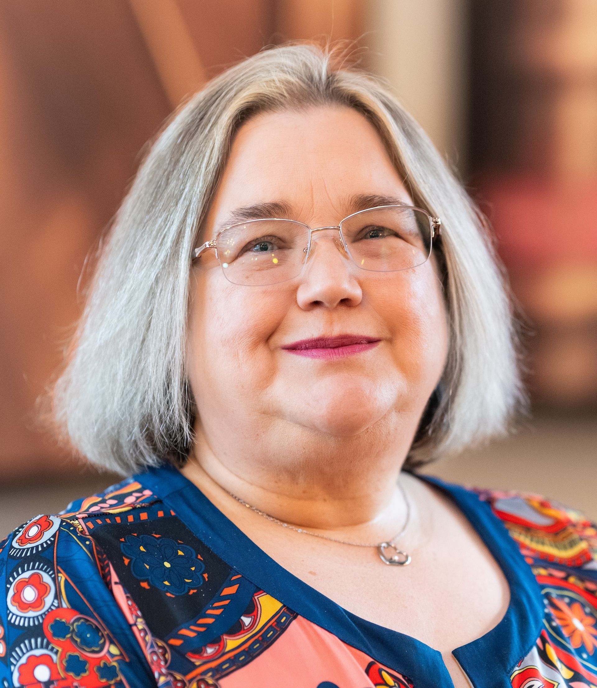 Portrait of a smiling person with glasses and shoulder-length gray hair, wearing a colorful patterned top.