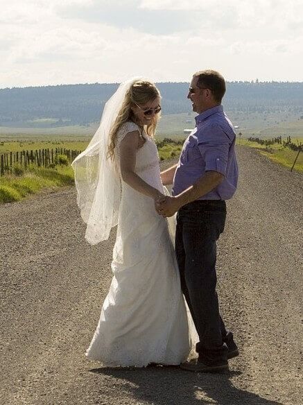 Bride and groom holding hands on a rural road, with fields and hills in the background.