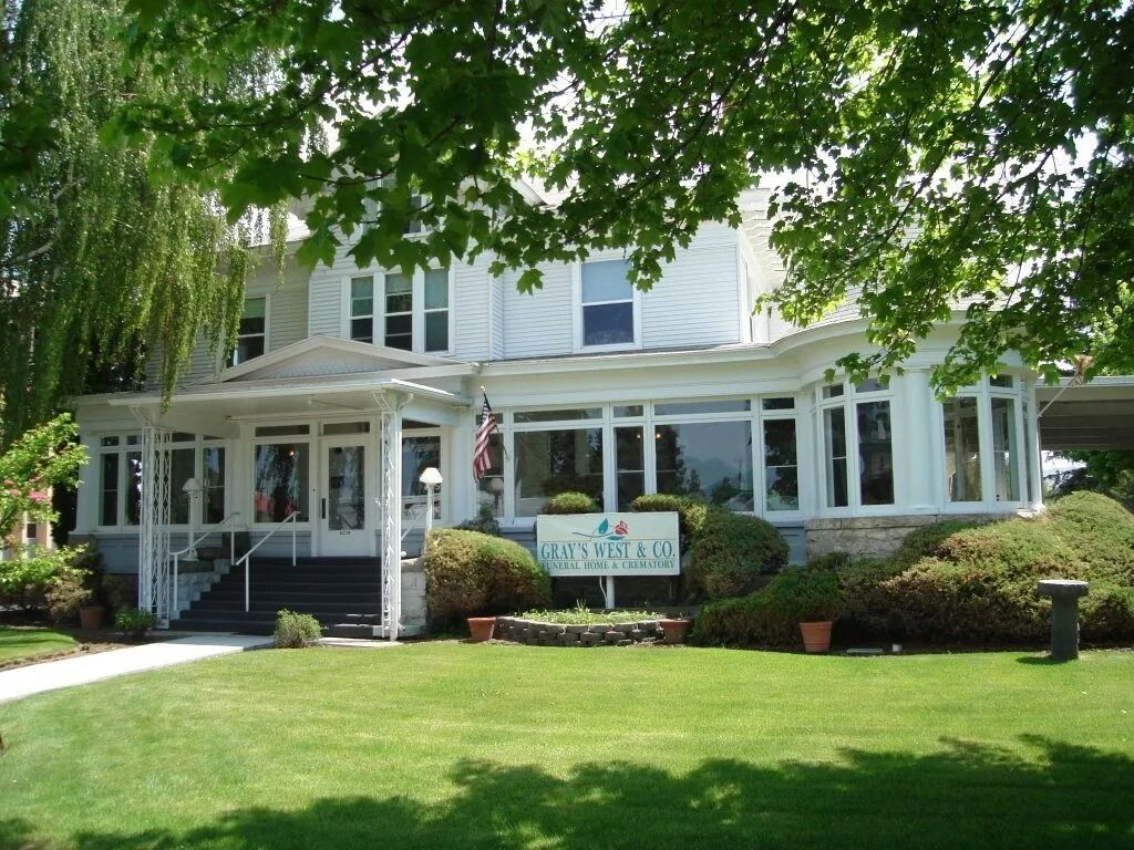 White house with porch and sign, shaded by trees, fronted by a green lawn